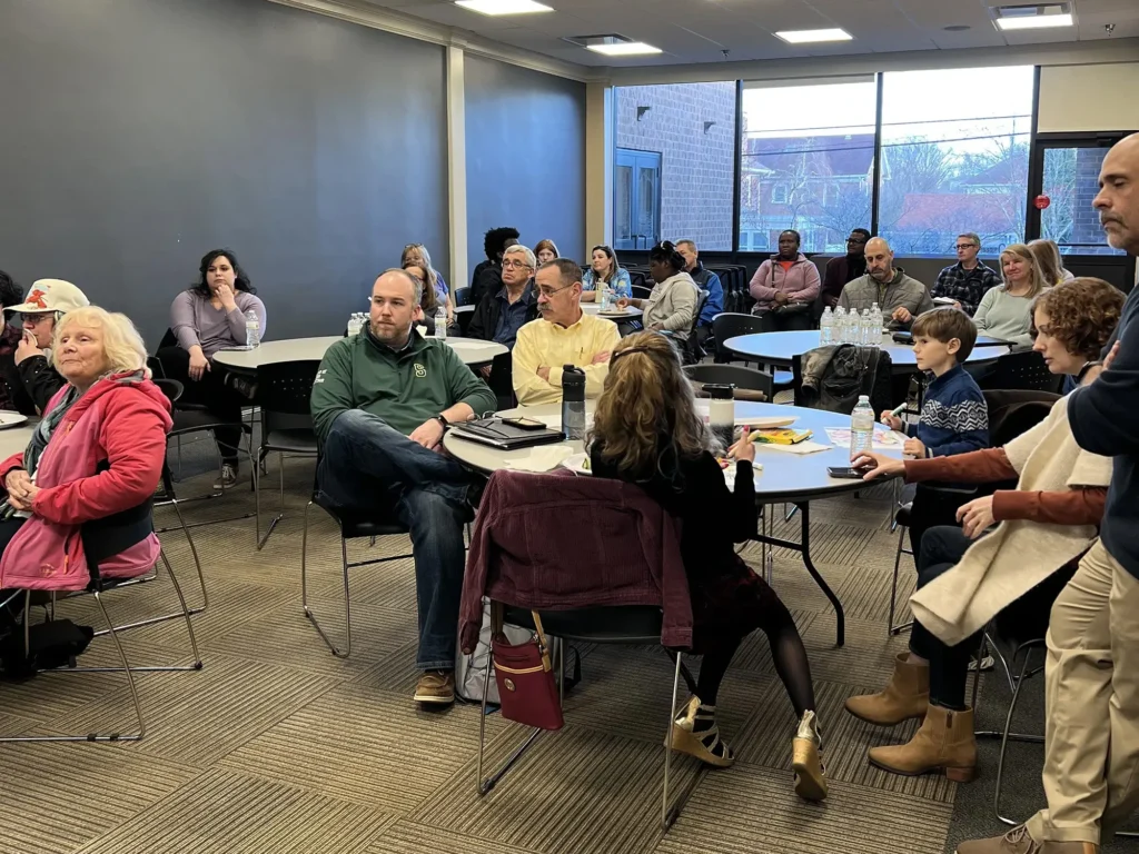 A large group of adults and a few children are seated in a conference room. They are arranged around several round tables, listening attentively to someone who is out of frame.